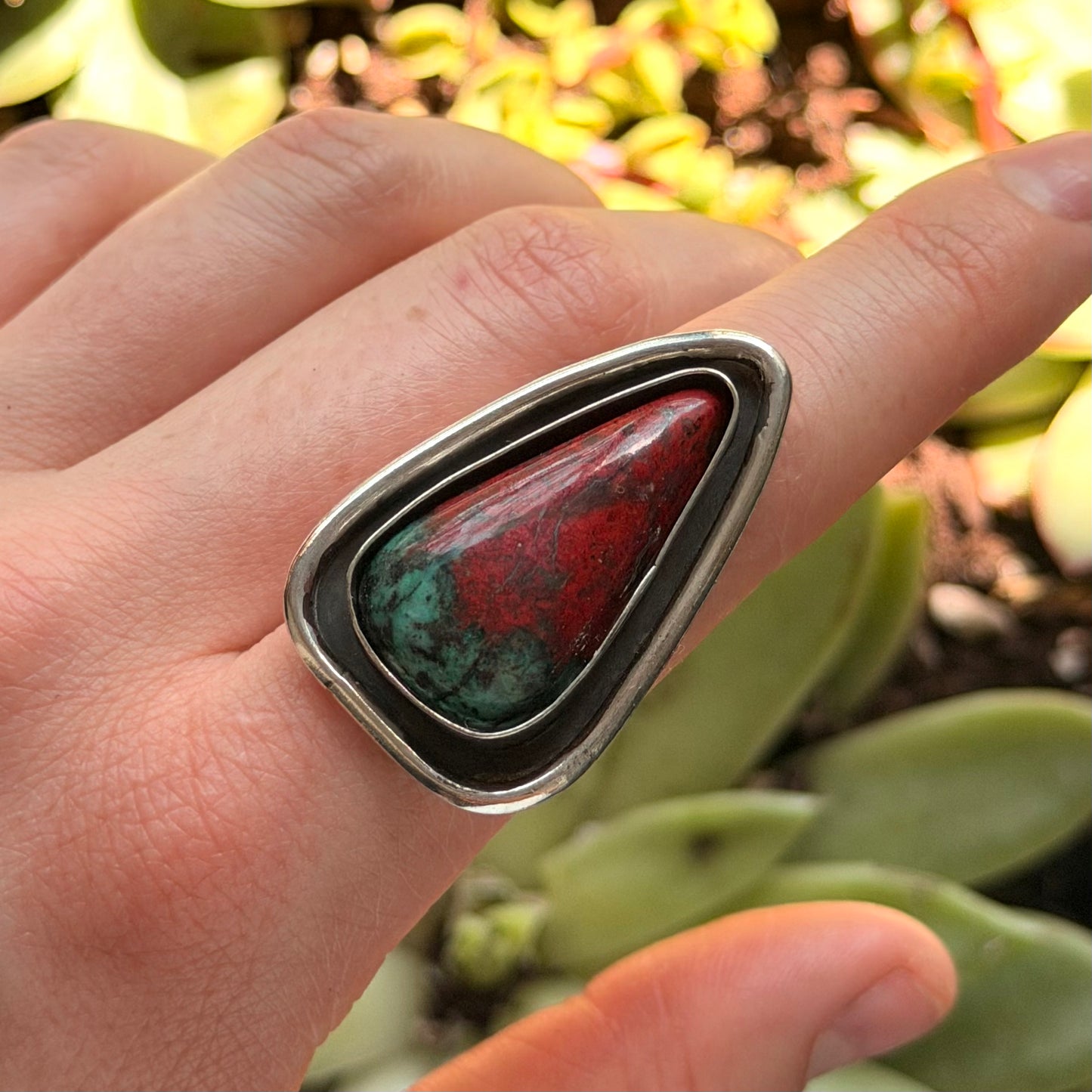 Hand wearing a ring with a red and green stone against a blurred green background