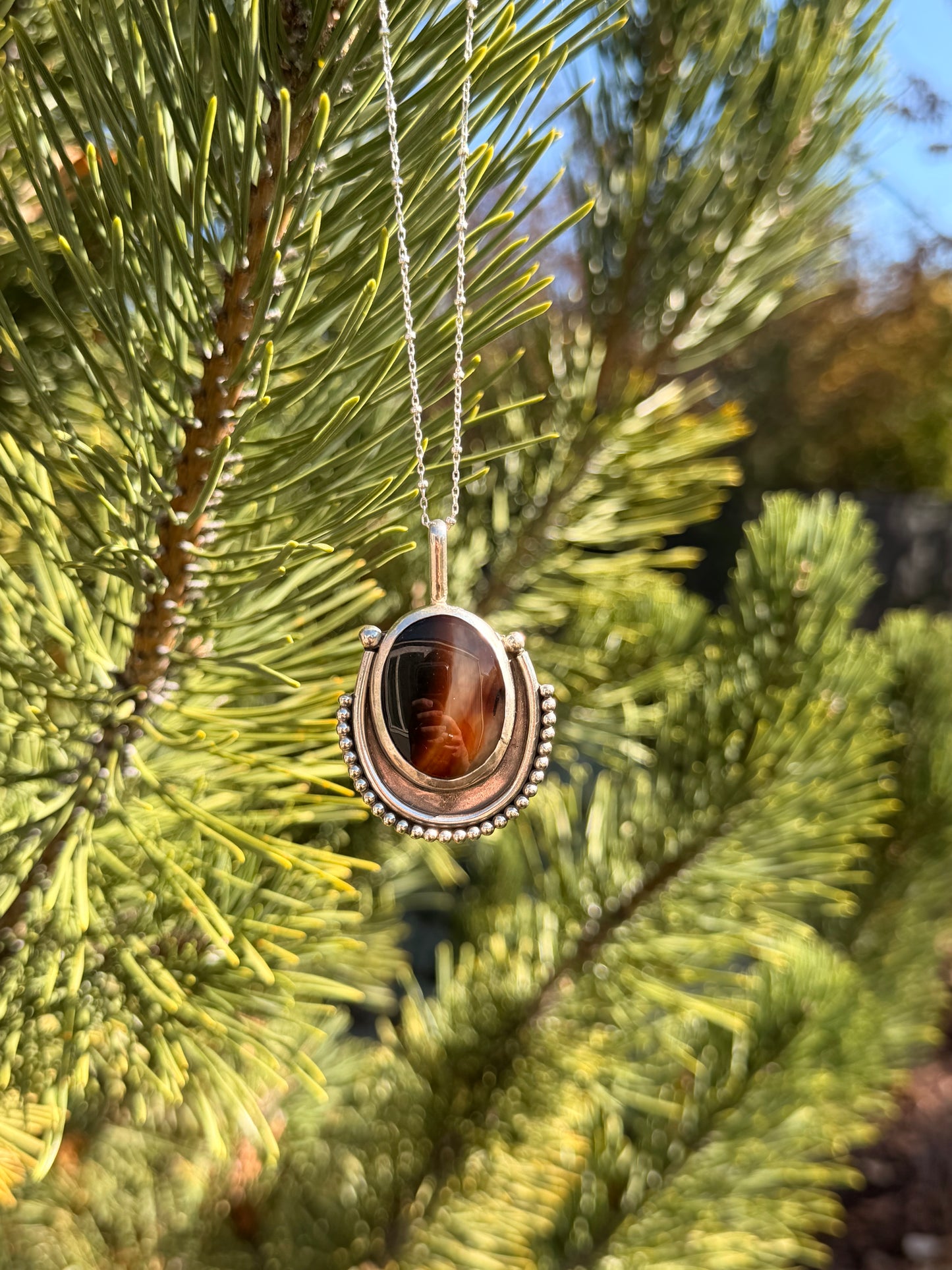 A silver pendant necklace with a polished agate stone hanging from a silver chain against a blurred background of green pine needles.