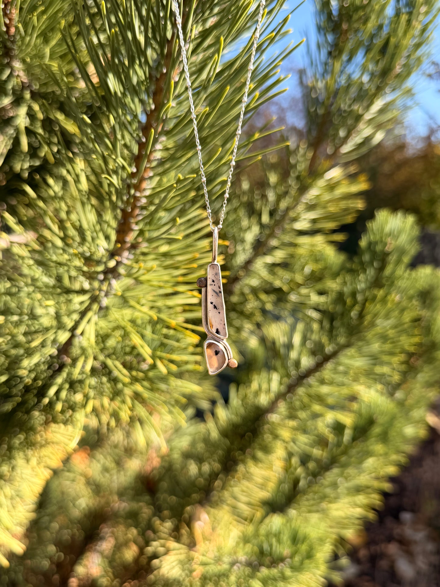 A silver pendant necklace with a polished agate stone hanging from a silver chain against a blurred background of green pine needles.