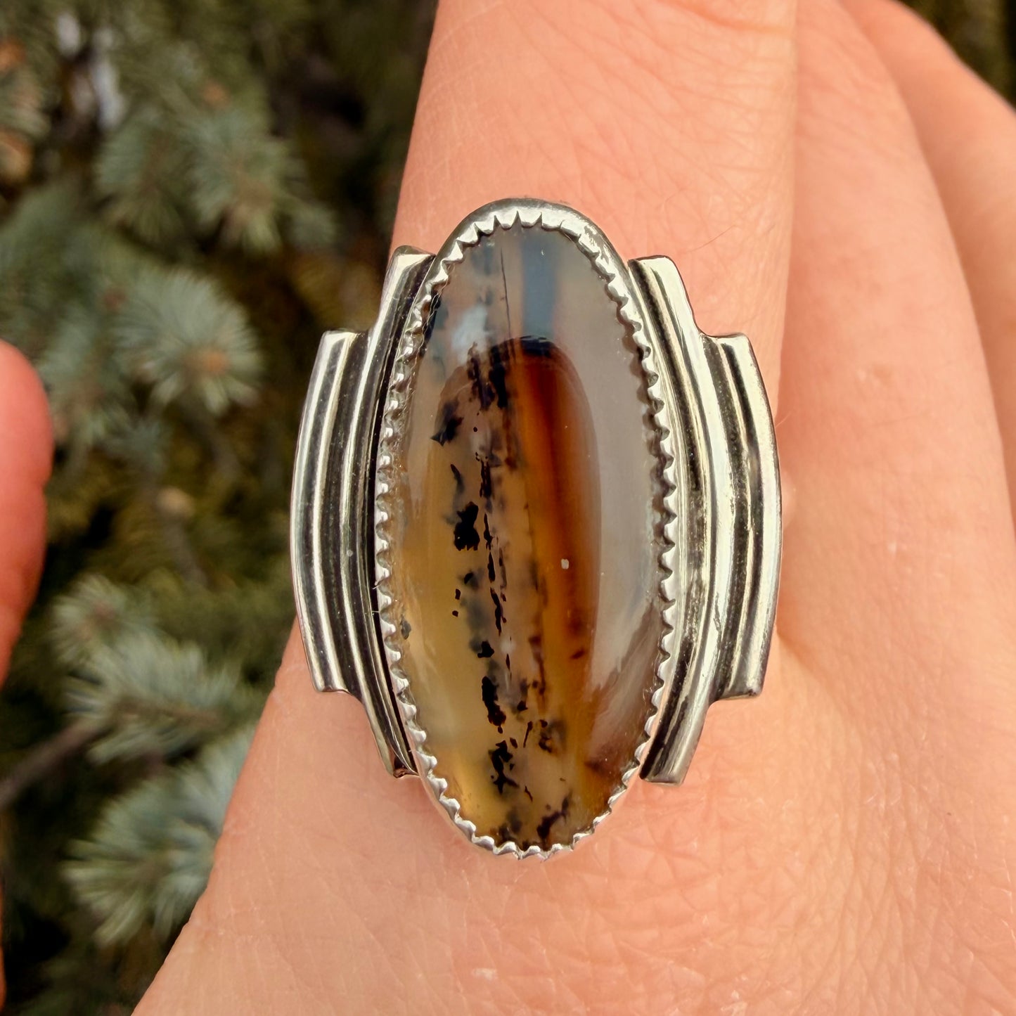 Silver ring with a large, oval-shaped gemstone on a person's finger with a blurred natural background.
