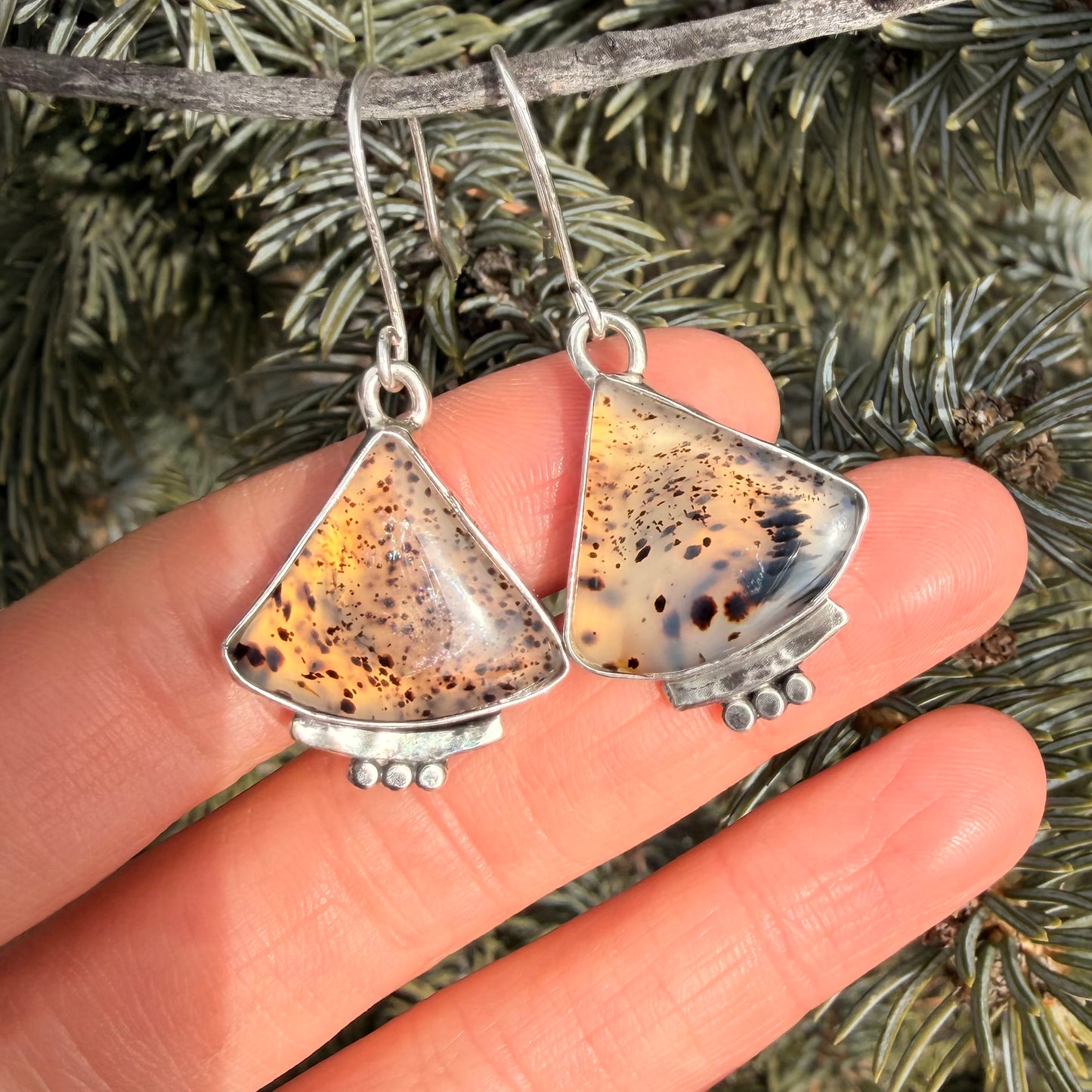 Triangular earrings with speckled design held between fingers against a natural background