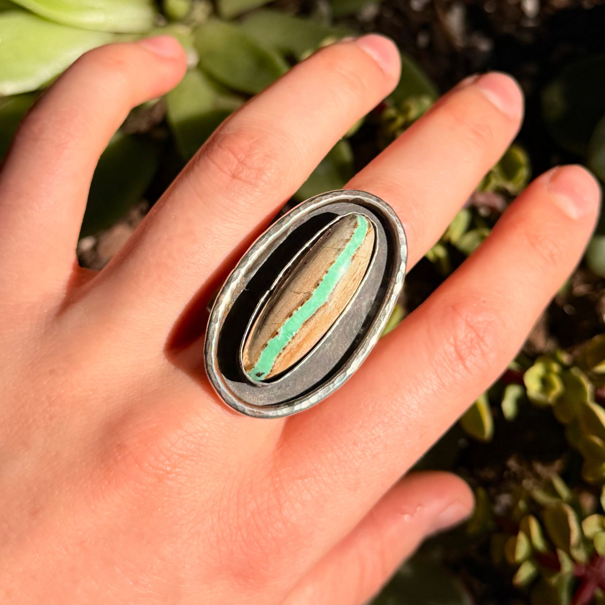 Hand wearing a ring with a shadow box surrounded by green foliage.