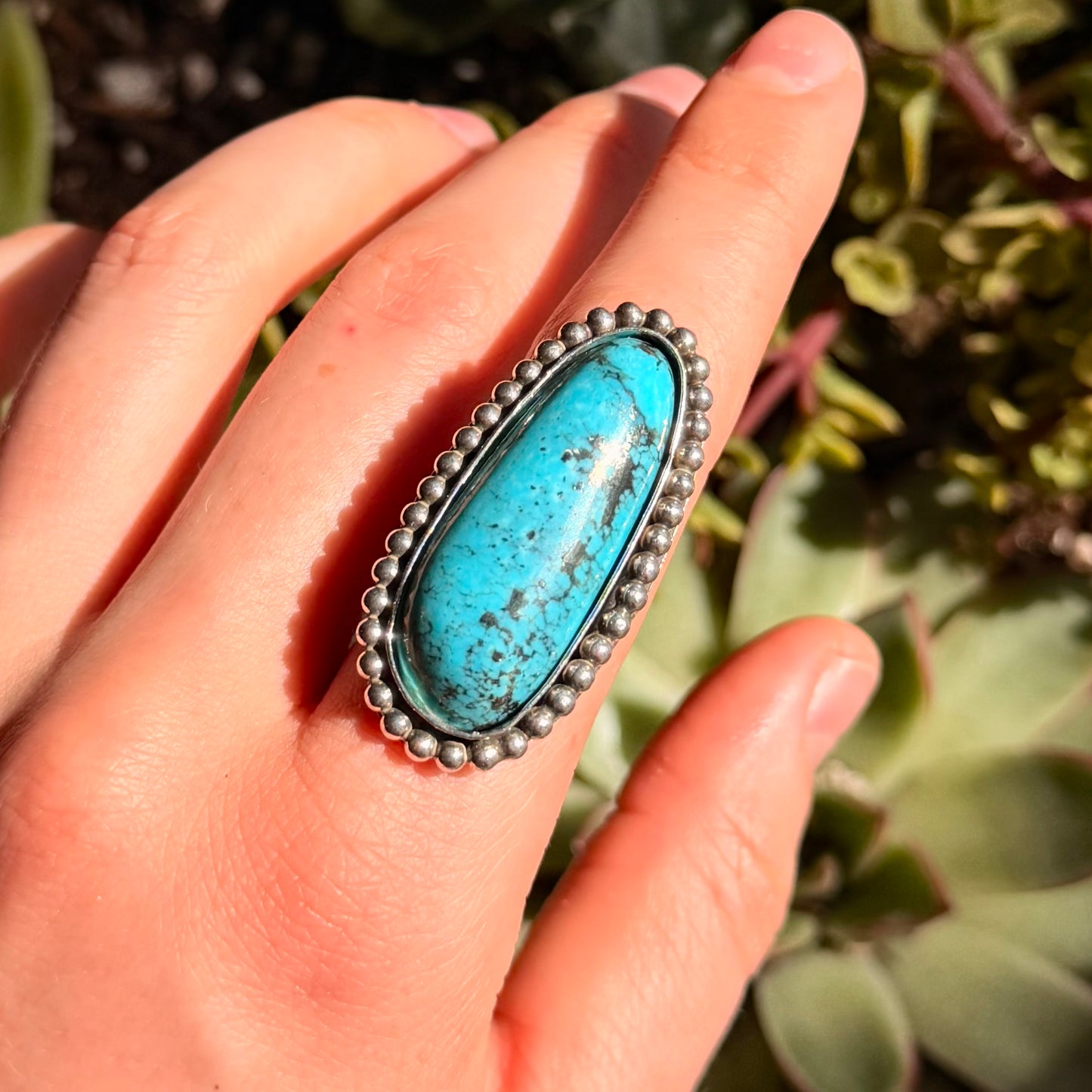 Turquoise ring with silver beaded wire on a hand in front of green plants