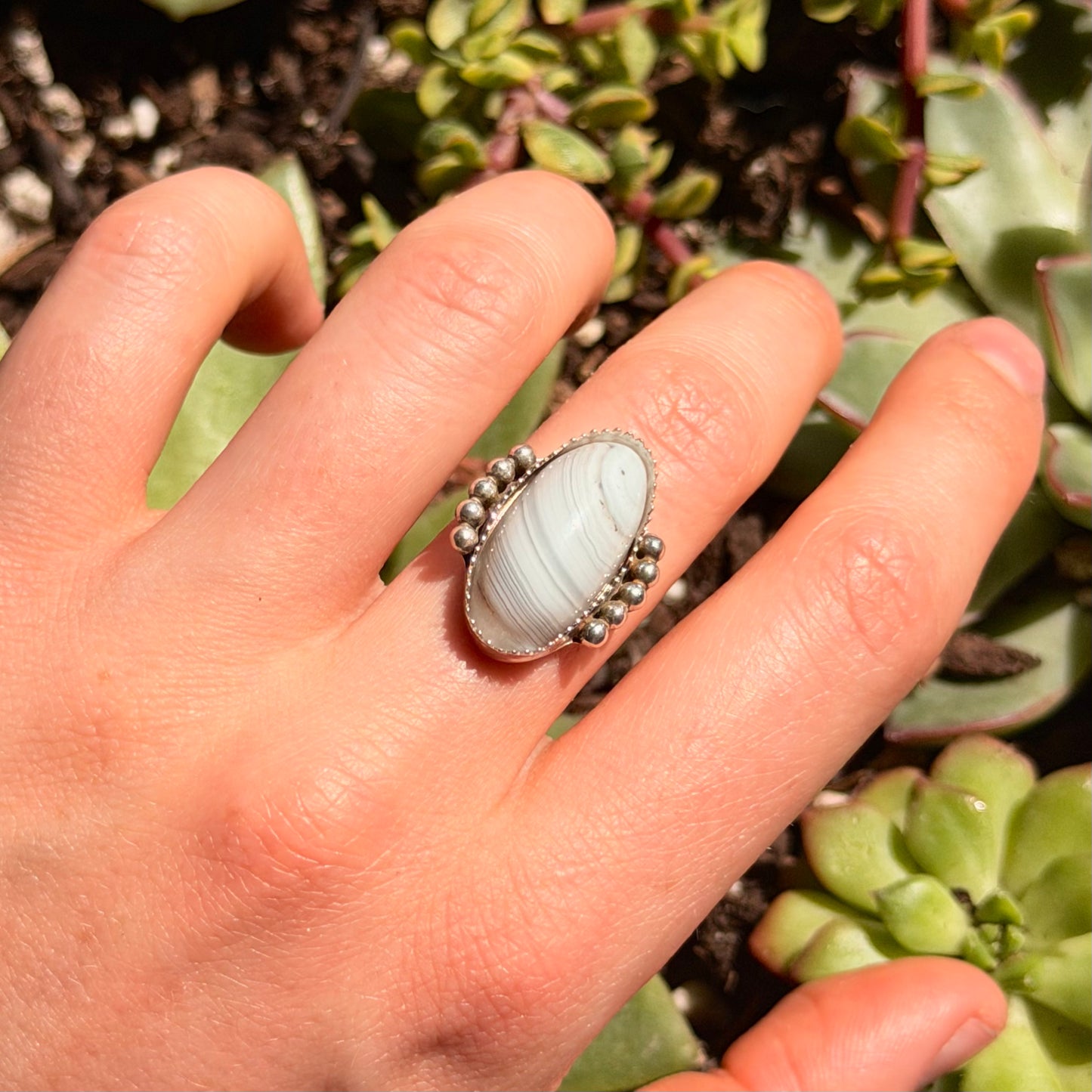Hand wearing a ring with a white stone against a background of green plants