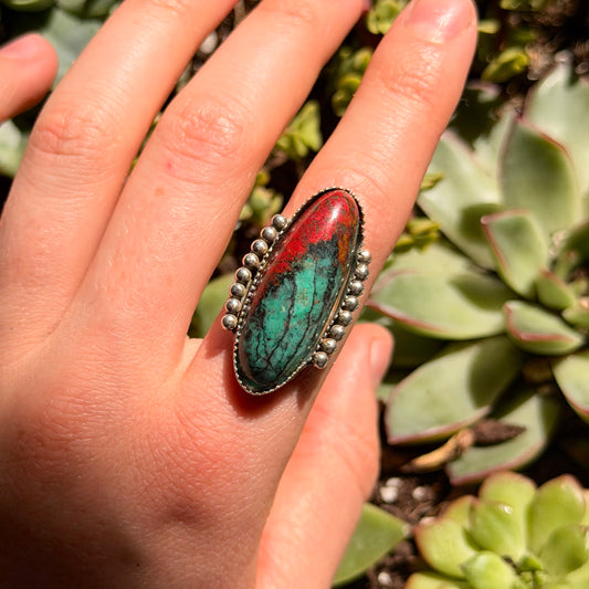 Red and green stone ring on finger with blurred green background