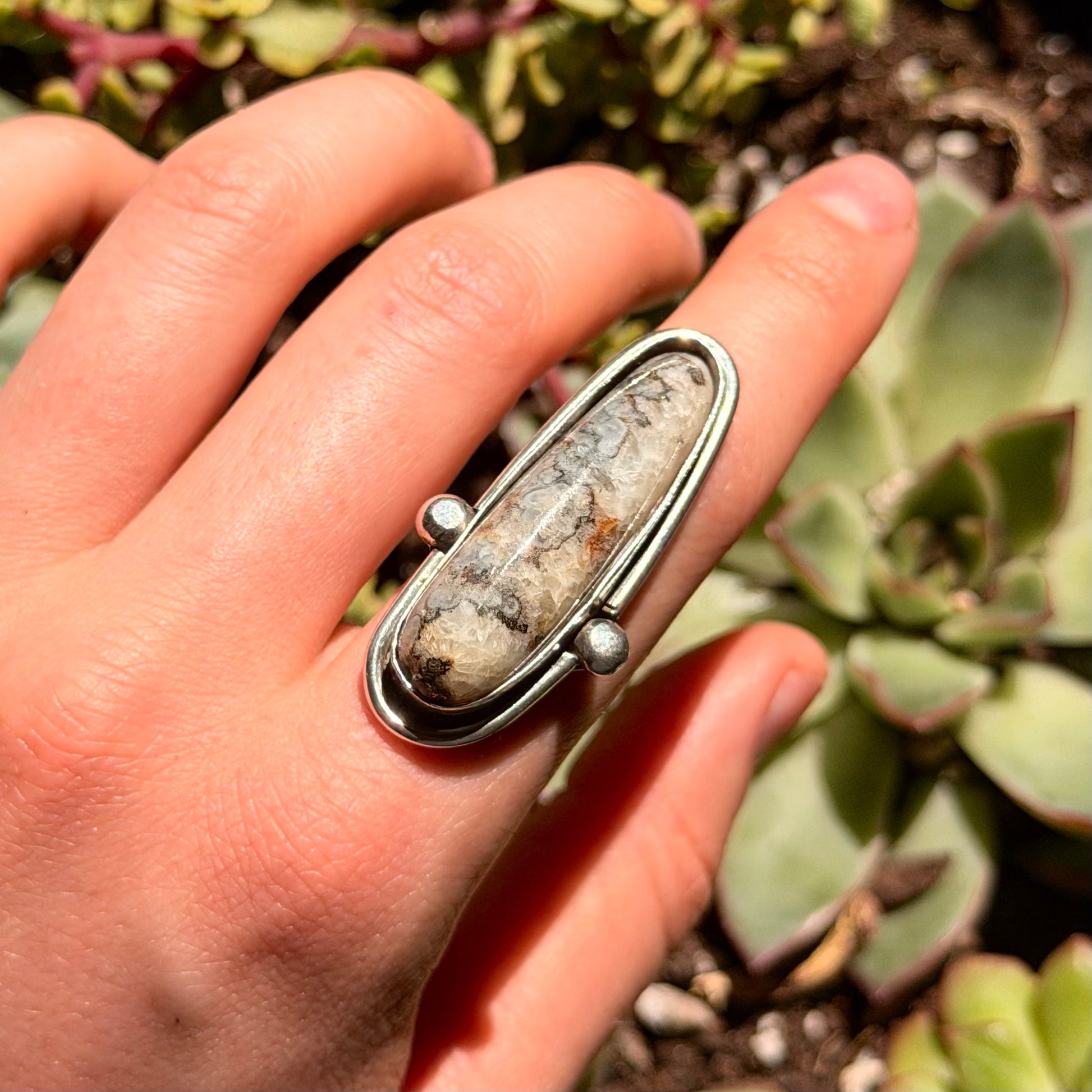 Grey and cream agate stone with pyrite streak surrounded by wire and balls ring without shank. Back ground is green plants