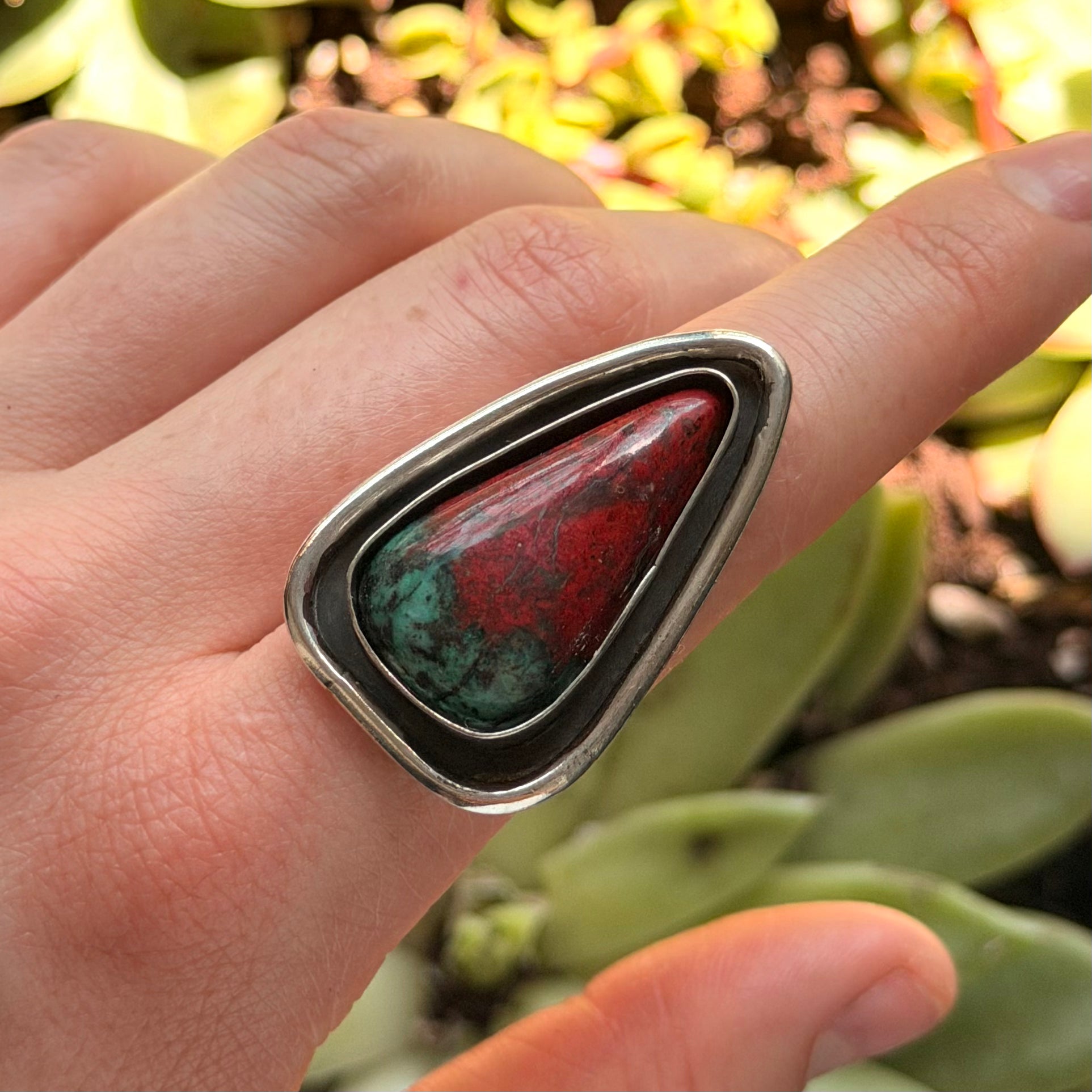 Hand wearing a ring with a red and green stone against a blurred green background