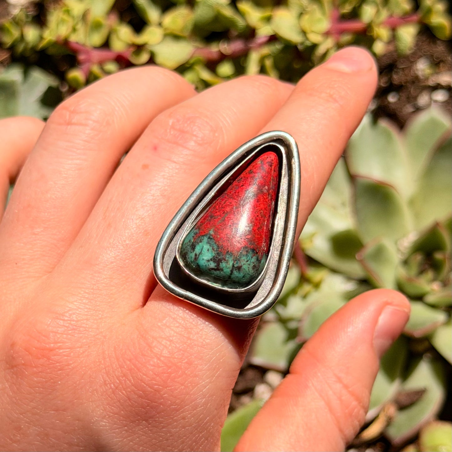 Ring with a red and green gemstone on a finger, with a natural background of greenery.