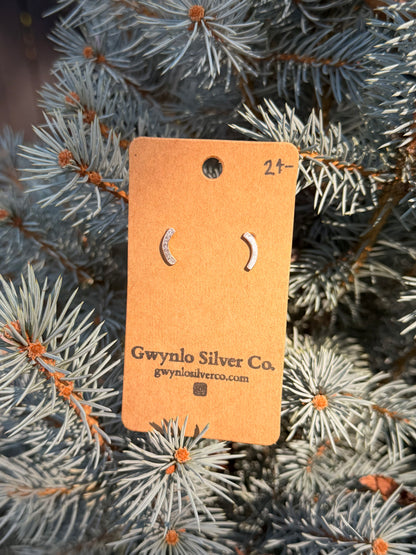 A pair of small crescent moon stud earrings in silver, displayed on a brown cardboard tag that reads 'Gwyno Silver Co.' and 'gwynosilverco.