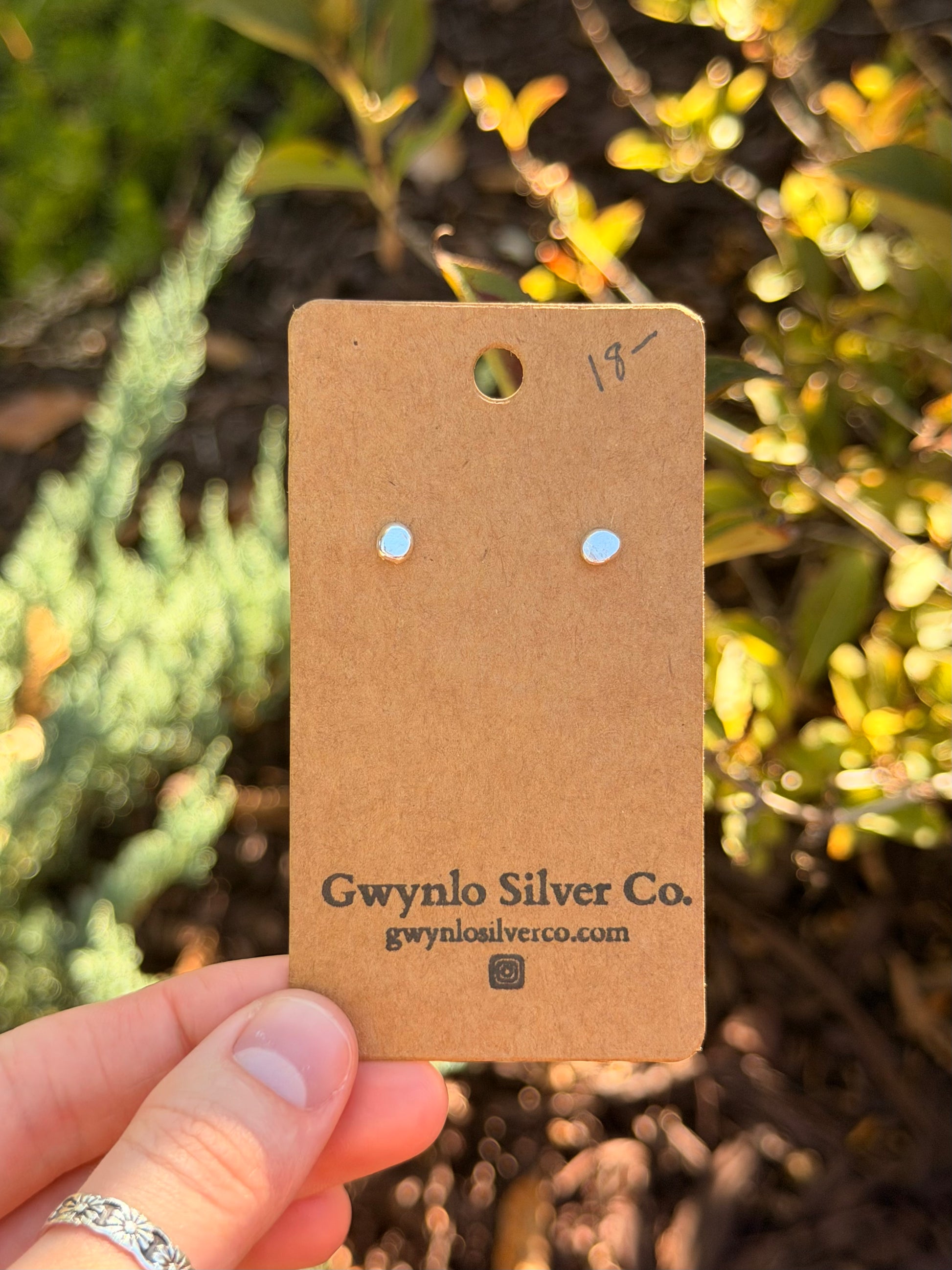 A hand holds a brown cardboard jewelry card displaying two small silver stud earrings set against a blurred outdoor garden background. The card is branded 'Gwynlo Silver Co.