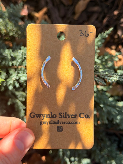 A pair of silver crescent-shaped earrings on a brown kraft display card held by a hand, with green foliage in the background.