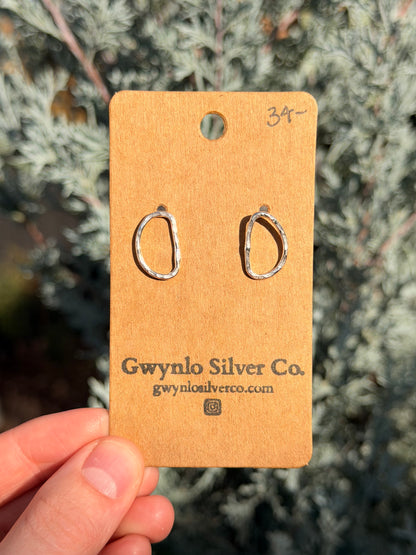 D Shaped hammered earrings on cardboard card held by hand with green foliage background.