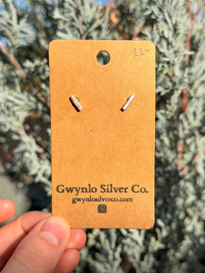 A pair of small silver stud earrings displayed on a tan cardboard backing card held by a hand, with blurred green foliage in the background.