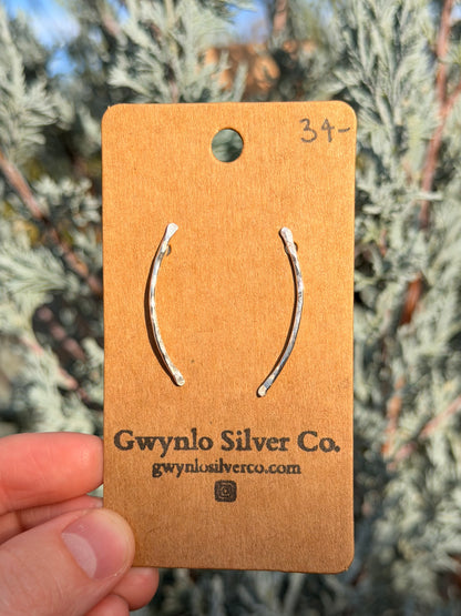 A pair of curved silver hoop earrings displayed on a brown  card held by a hand, with blurred green foliage in the background. The card is branded 'Gwynlo Silver Co.