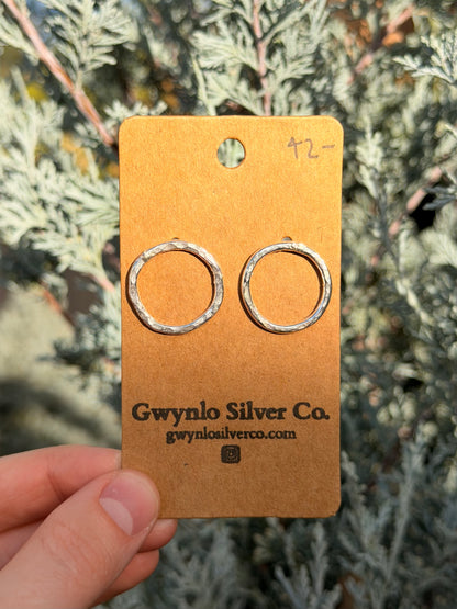 A pair of textured silver hoop earrings displayed on a brown kraft card held by a hand, with green foliage in the background.