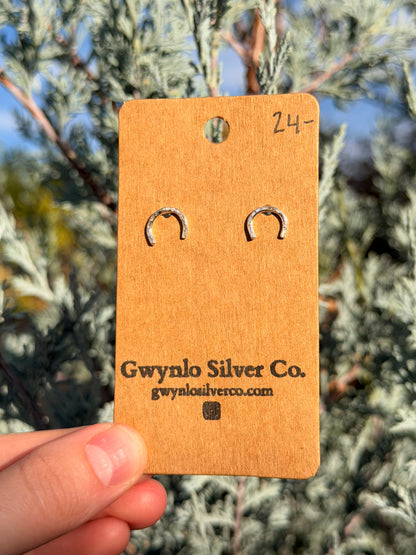 A pair of small silver horse shoe shaped hoop earrings on a brown cardboard display card, held by a hand in front of blurred green foliage.