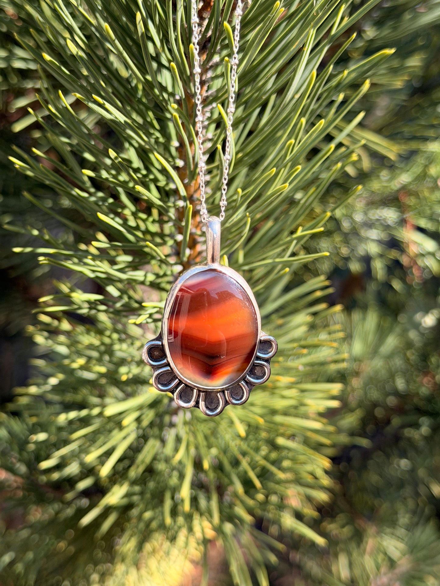 A silver pendant necklace with a polished agate stone hanging from a silver chain against a blurred background of green pine needles.