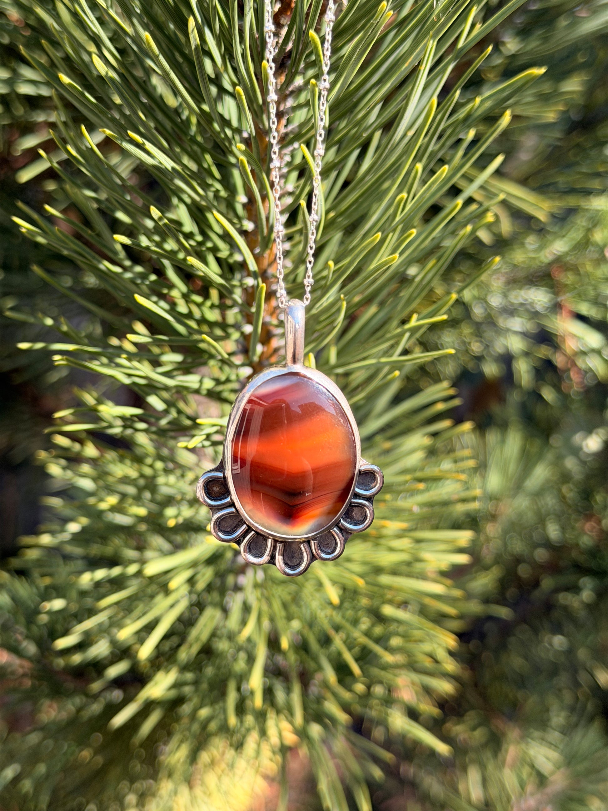 A silver pendant necklace with a polished agate stone hanging from a silver chain against a blurred background of green pine needles.