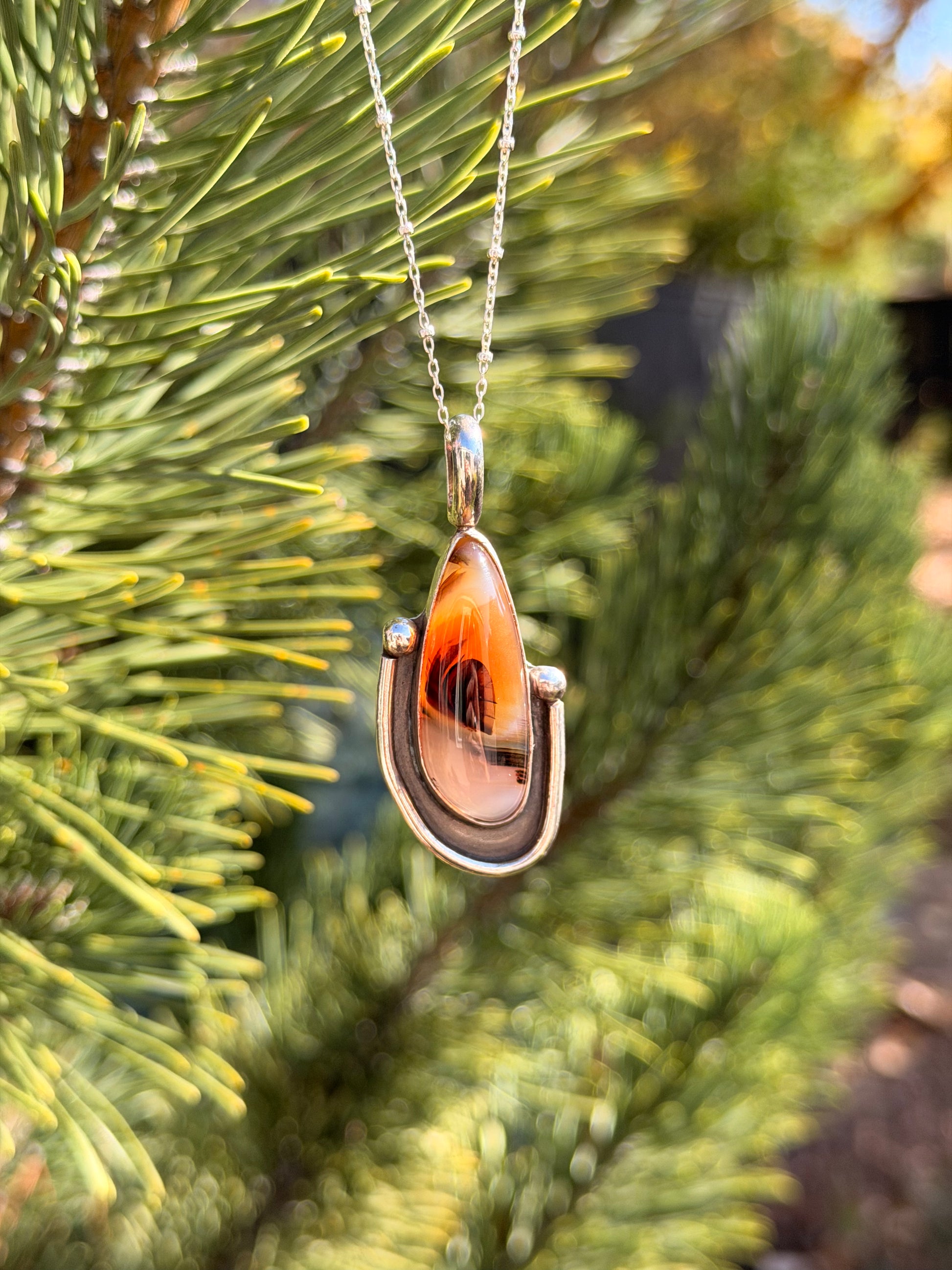 A silver pendant necklace with a polished agate stone hanging from a silver chain against a blurred background of green pine needles.