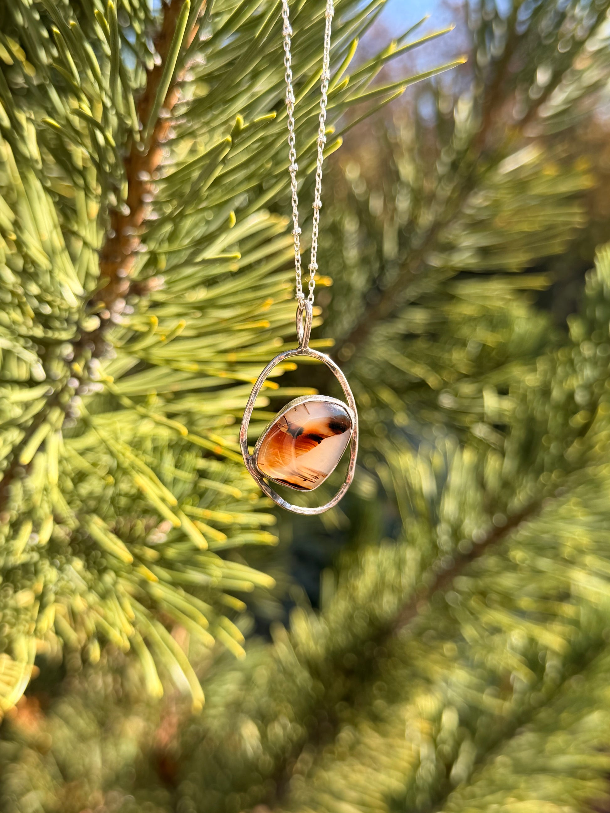 A silver pendant necklace with a polished agate stone hanging from a silver chain against a blurred background of green pine needles.