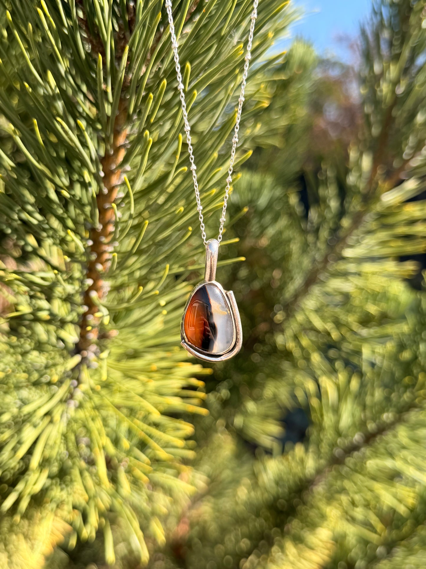 A silver pendant necklace with a polished agate stone hanging from a silver chain against a blurred background of green pine needles.