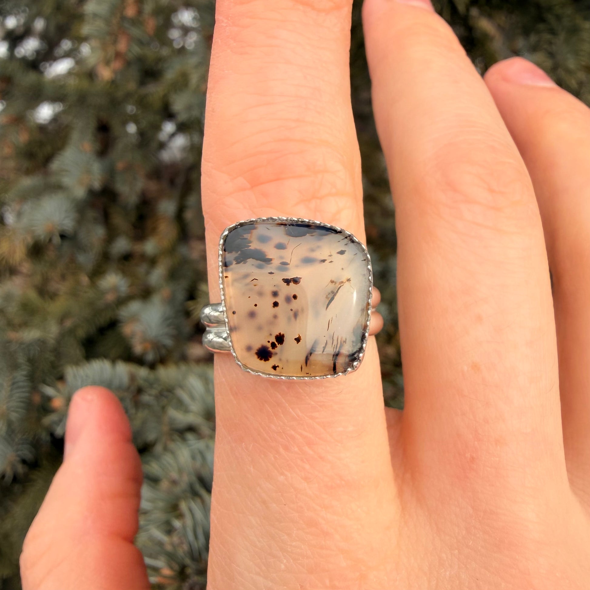 Hand wearing a ring with a speckled stone, blurred greenery in the background
