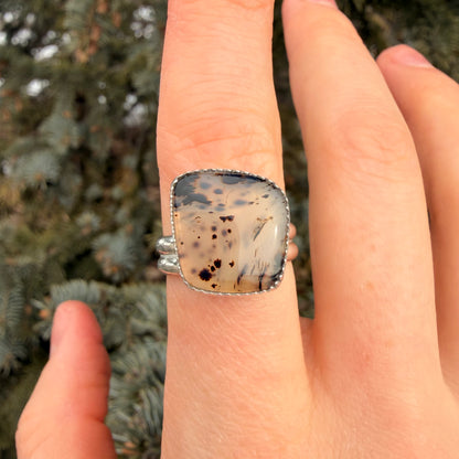 Hand wearing a ring with a speckled stone, blurred greenery in the background