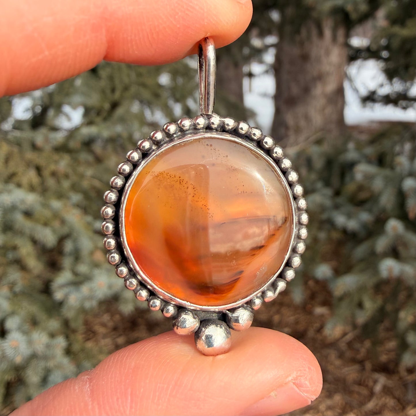 Silver pendant with an orange stone held between two fingers against a blurred natural background