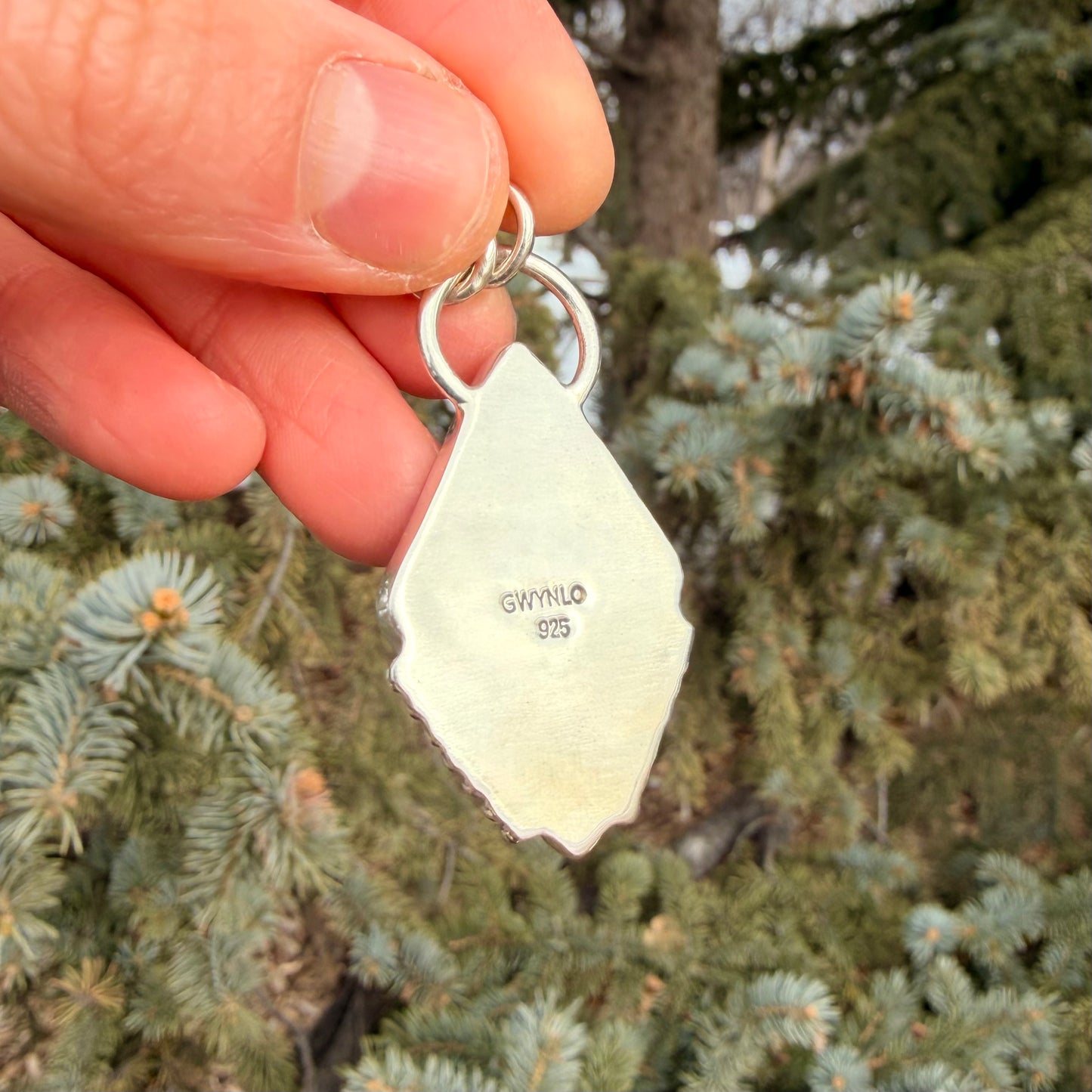 Hand holding a silver pendant with 'Gwynlo' stamp against a green foliage background