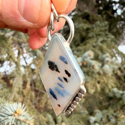 Decorative pendant with blue and black designs held against a blurred natural background