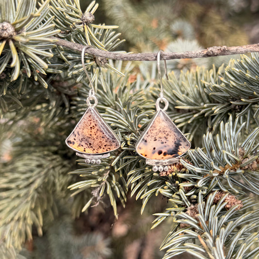 Pair of triangle-shaped earrings with a speckled design hanging on a branch of a pine tree.