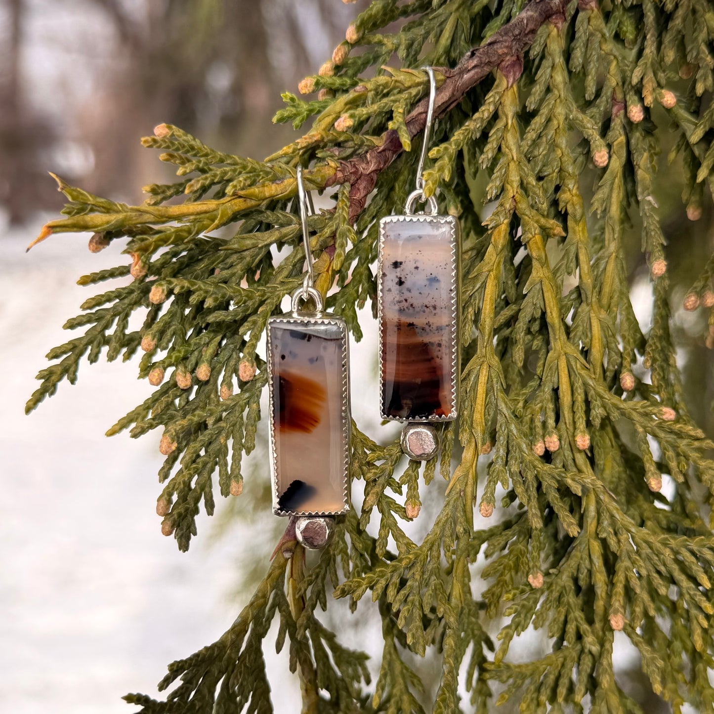 Pair of earrings with a rectangular design hanging on a branch with green leaves.